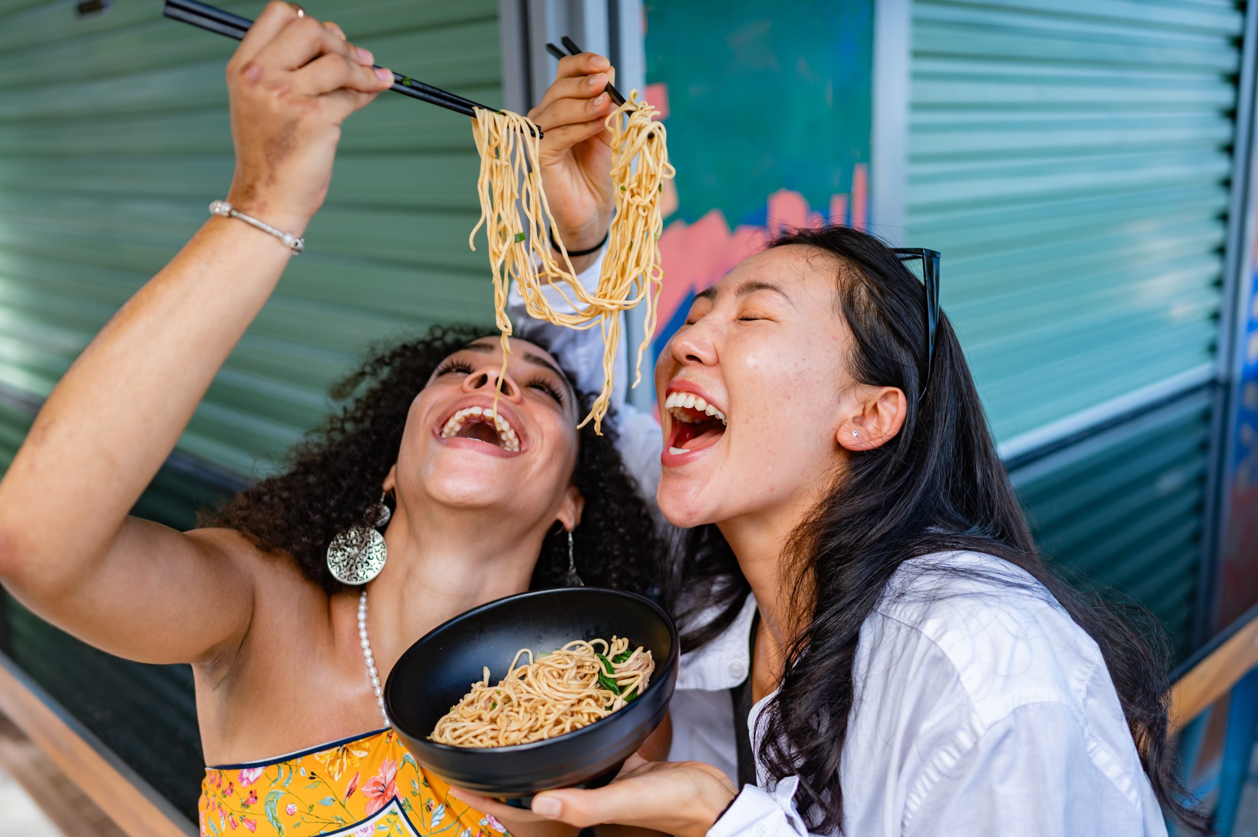 Dois amigas comendo macarrão juntos, sorrindo e aproveitando uma refeição descontraída e divertida.