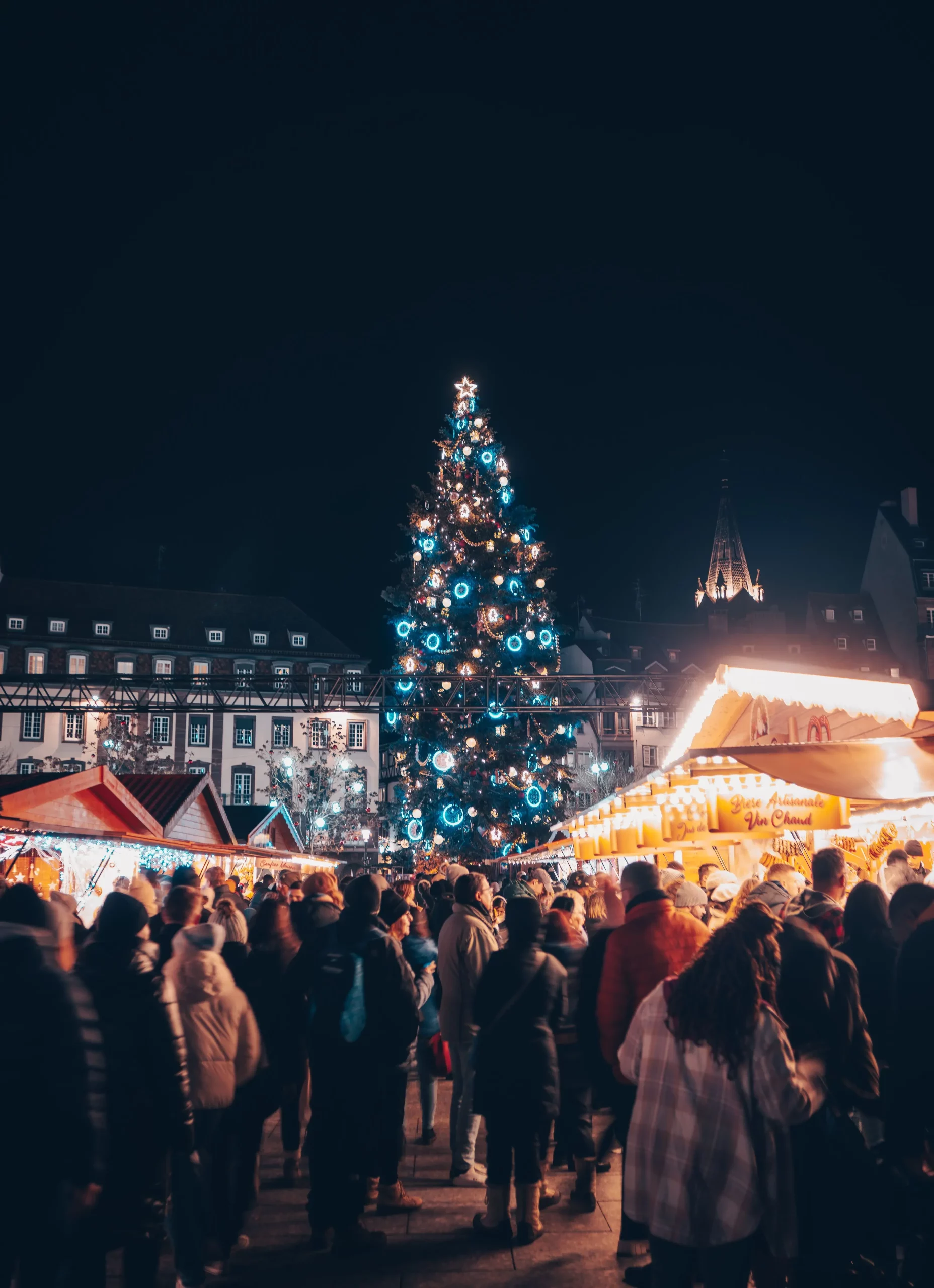 Um movimentado mercado de Natal em Estrasburgo à noite, com uma enorme árvore de Natal iluminada com luzes azuis e brancas, rodeada por bancas tradicionais de madeira e visitantes agasalhados.